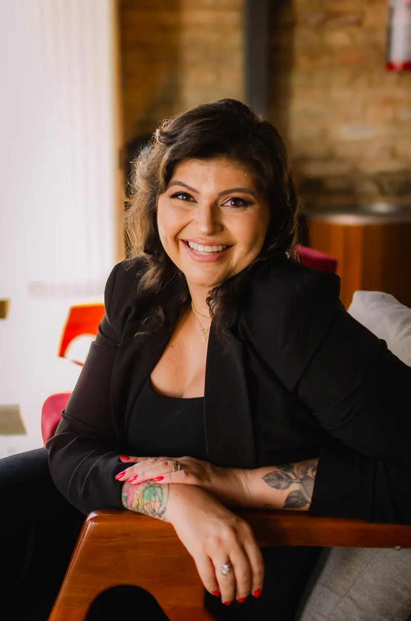 Retrato de Kelly Pinheiro sorrindo para a câmera, de roupa preta, sentada em uma poltrona de madeira